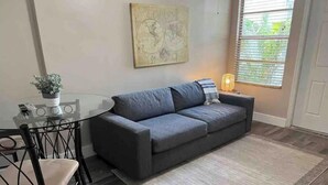 Un salon moderne avec un canapé gris, une table en verre et un fauteuil. Il y a une carte accrochée au mur et une fenêtre avec des stores.