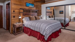 Un dormitorio con cabecera de madera, cama con ropa de cama a rayas, falda para cama roja y mesita de noche con una lámpara, además de un espejo que refleja la habitación.