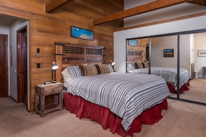 Een slaapkamer met een houten hoofdbord, een bed met gestreepte beddengoed, een rode bedrok, een nachtkastje met een lamp en een spiegel die de kamer reflecteert.