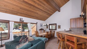 Una sala de estar acogedora con sofá, mesa de comedor y sillas. Hay una zona de cocina con armarios de madera y una lámpara colgante.