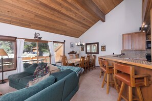 Een gezellige woonkamer met een bank, een eettafel en stoelen. Er is een keuken met houten kastjes en een hangende lamp.