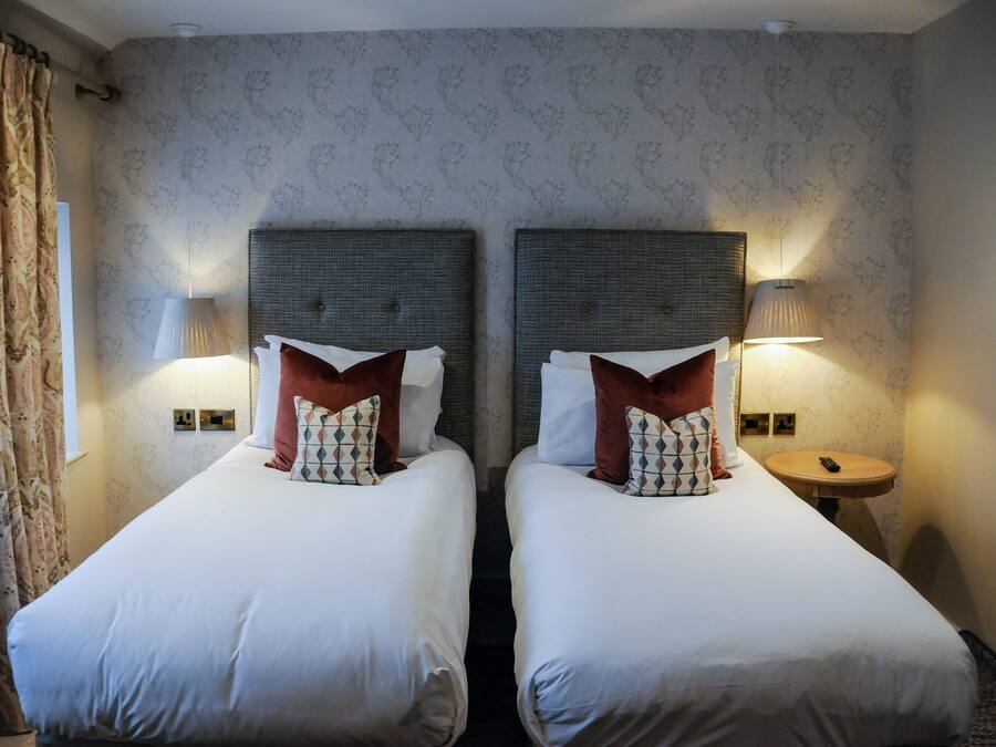 A hotel room with two beds, each with a headboard, white bedding, and patterned throw pillows. There are wall-mounted lamps on either side of the beds.