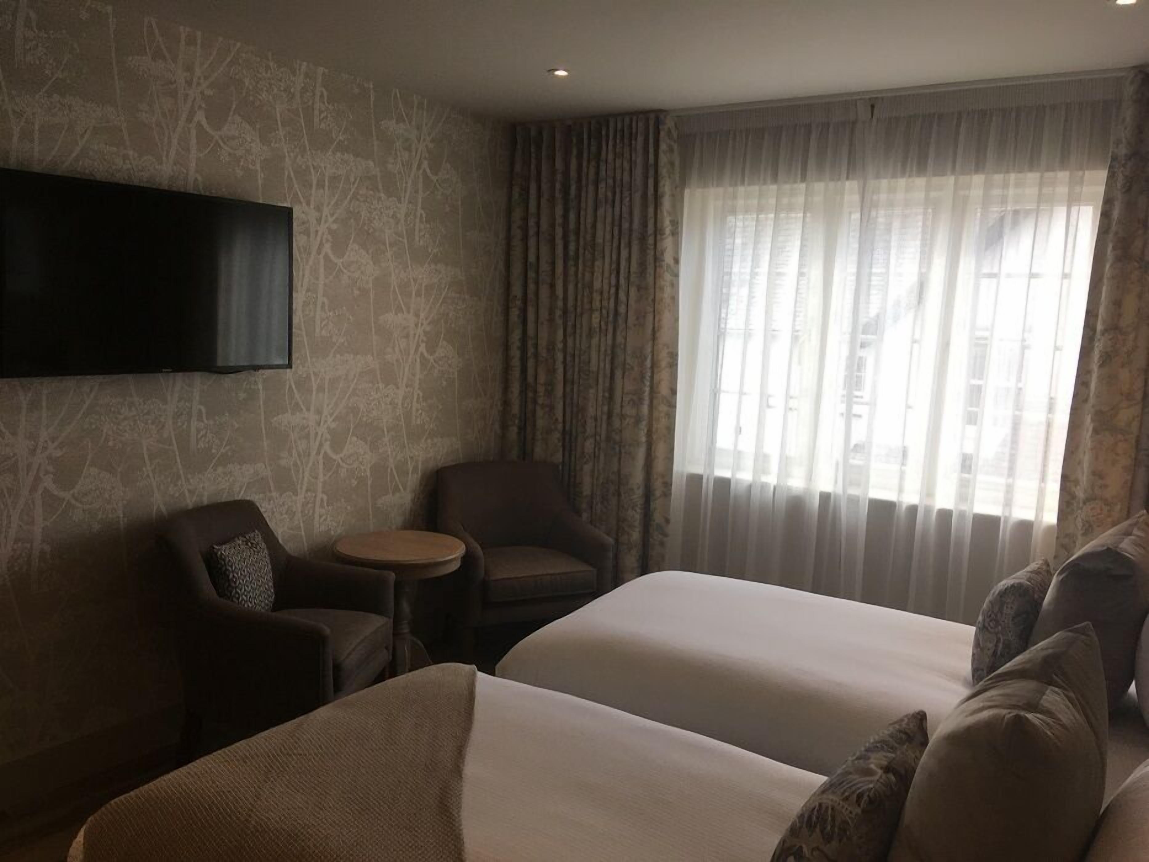 A hotel room with two beds, a TV mounted on the wall, a small round table, a chair, and a window with sheer curtains.