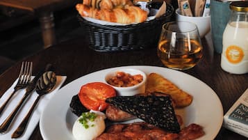Une assiette de petit-déjeuner avec des œufs, du bacon, des toasts, des haricots et une tomate. Il y a des croissants dans un panier, un verre de bière et une bouteille de lait sur la table.