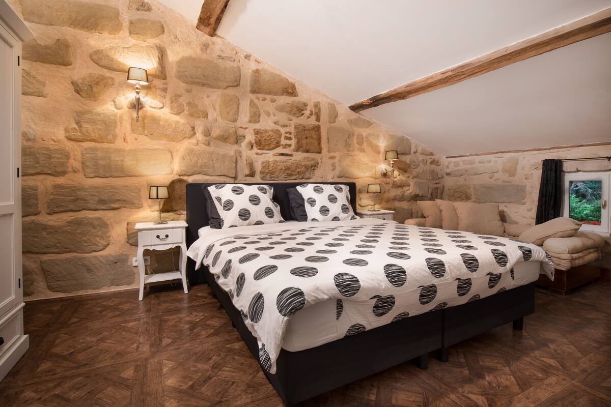 A bedroom with a stone wall, a bed with a patterned bedspread, two bedside tables with lamps, and a wooden ceiling beam.
