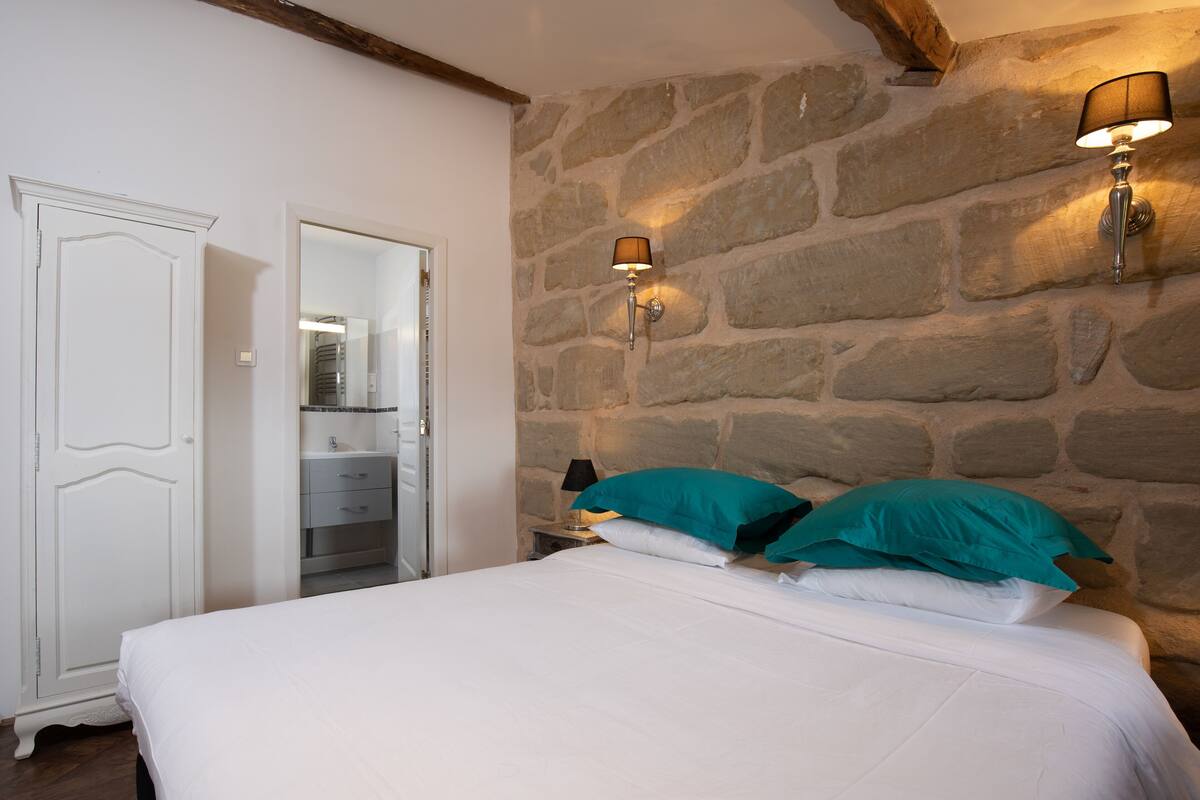 A bedroom with a stone wall, a bed with white bedding, teal pillows, and two wall-mounted lamps.