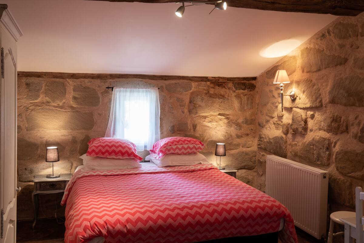 A bedroom with a stone wall, a bed with pink bedding, two bedside lamps, a window with a white curtain, and a white radiator.