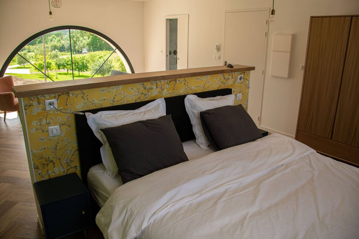 A bedroom with a bed, a nightstand, a wardrobe, and a view of greenery through a window.