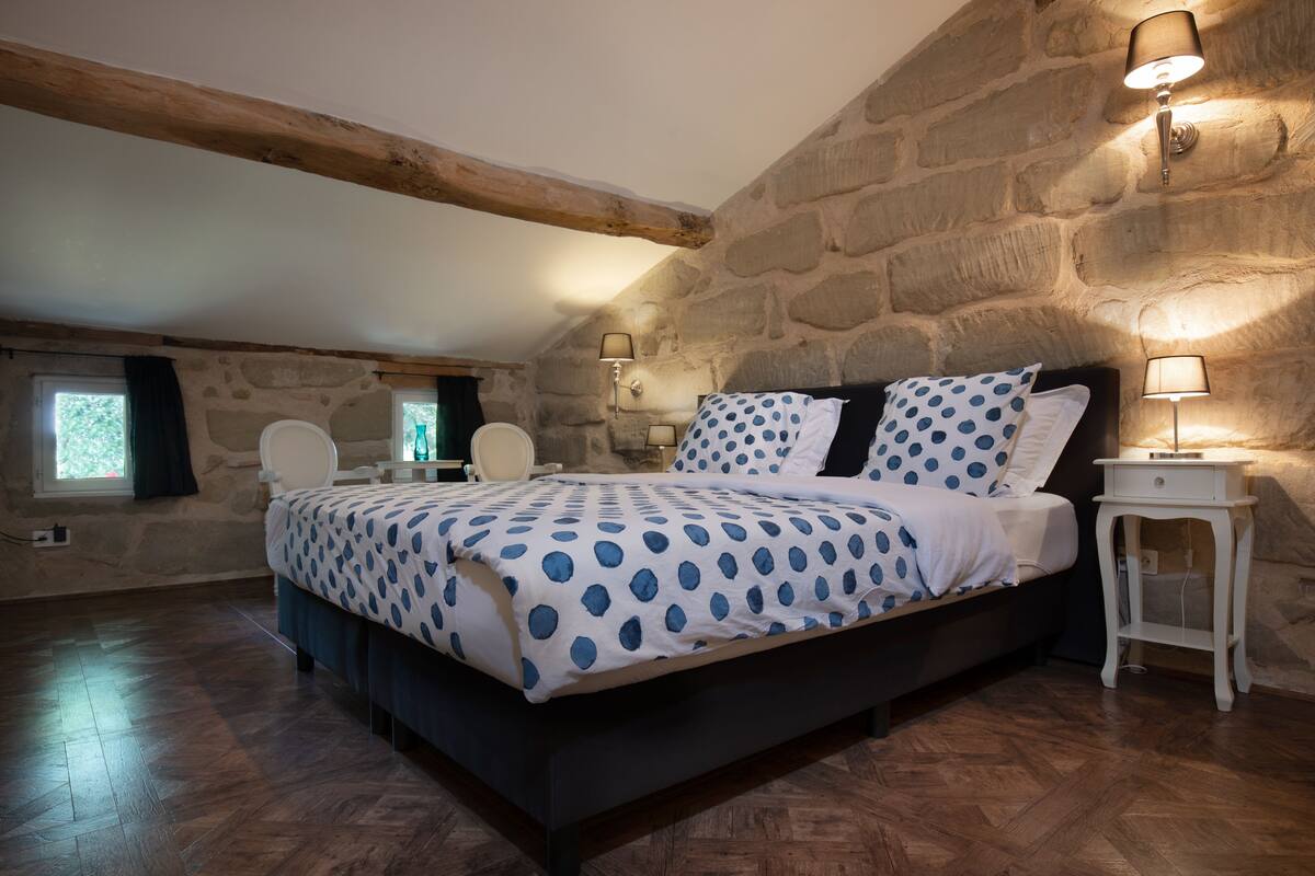A bedroom with a bed, two bedside tables, a chair, a window, and stone walls.