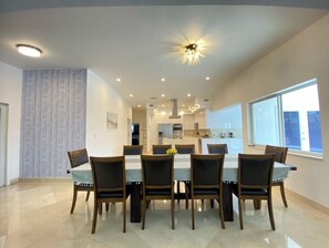A spacious dining area with a long table and chairs, a modern kitchen in the background, and large windows providing natural light.