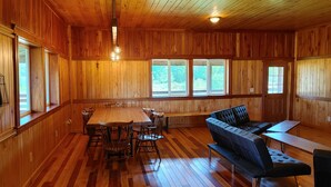 Ein geräumiges Zimmer mit Holzwänden und Holzdielen, ein großer Esstisch mit Stühlen, eine Couch und ein kleiner Tisch.