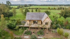 Une maison en bois avec un toit en pente, entourée d’un jardin bien entretenu et, au loin, d’un grand champ verdoyant.