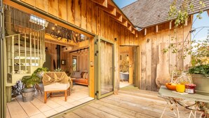 Une cabane en bois avec une entrée dotée d’une porte en verre, un coin salon confortable avec un canapé et des fauteuils, et une table à manger agrémentée d’un panier et de fruits.