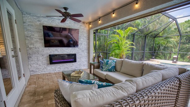 A cozy indoor patio with a white brick wall, a flat-screen TV mounted above a fireplace, wicker furniture, and a view of lush greenery through large windows.
