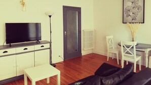 Una sala de estar con un televisor sobre un mueble, una mesa blanca con sillas, un sofá negro y una puerta.