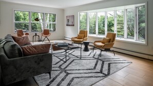 A spacious living room with a modern sofa, two armchairs, a coffee table, and a rug. Large windows offer a view of trees. - Birch Hill - Luxury Mountain Retreat (Windham)
