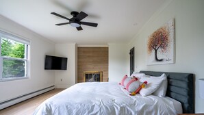 A modern bedroom with a large bed, a ceiling fan, a flat-screen TV, and a fireplace. - Birch Hill - Luxury Mountain Retreat (Windham)