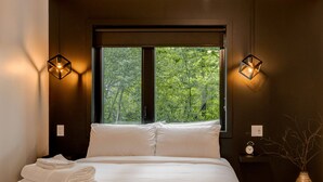 A bedroom with a large window, a bed with white linens, two wall-mounted lamps, and a bedside table with a clock and a vase. - The Experience SÔ Lodge (SHEFFORD)