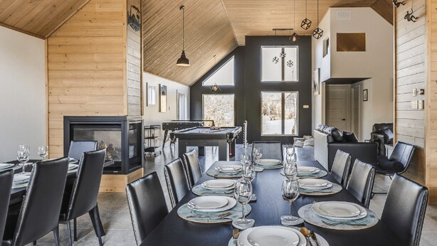 Un espace repas avec une longue table dressée pour six personnes, une cheminée et une vue sur l’extérieur.