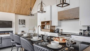 A modern kitchen with a dining area, featuring a central island, stainless steel appliances, and pendant lighting. - The Experience SÔ Lodge (SHEFFORD)