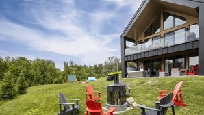 A modern house with a large deck, outdoor seating, and a fire pit. - The Experience SÔ Lodge (SHEFFORD)