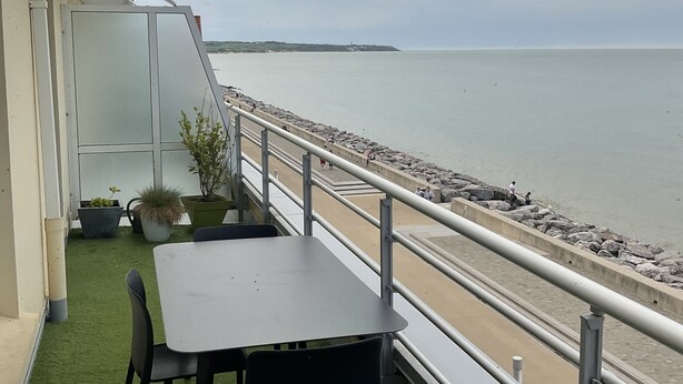 Une terrasse avec une table et des chaises, donnant sur une plage bordée d’une côte rocheuse et sur une île située au loin.