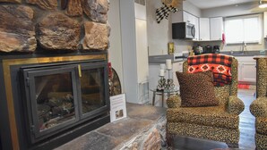 Una chimenea de piedra con protector de vidrio, un sillón estampado de leopardo y una cocina con armarios blancos y microondas.