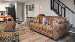 Una sala de estar con un sofá de cuero marrón, un sillón estampado de leopardo y un sillón con estampado. Se observa una zona de cocina con un refrigerador negro y una mesa de comedor con mantel a cuadros rojo y negro. En el fondo, se ve una escalera con barandilla negra.