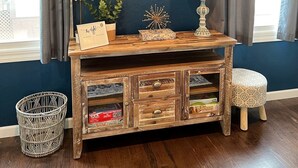 A room with a dark blue wall, a mounted flat-screen TV, a wooden console table with drawers, a small stool, and a wicker basket.