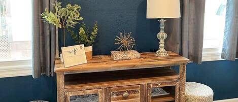 A room with a dark blue wall, a mounted flat-screen TV, a wooden console table with drawers, a small stool, and a wicker basket.