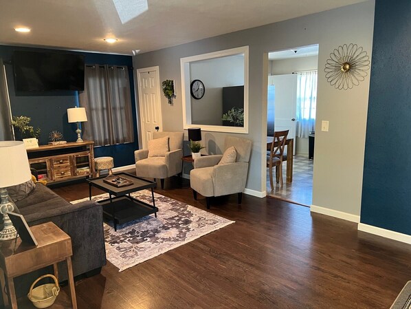 Un salon avec un parquet en bois foncé, un canapé gris, un fauteuil beige, une table basse et un téléviseur.