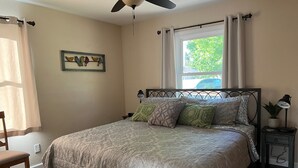 A bedroom with a bed, a ceiling fan, a window with curtains, a nightstand with a lamp, and a chair.