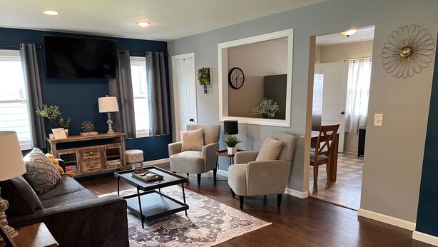 A living room with a dark blue accent wall, a flat-screen TV, a sofa, a coffee table, and a dining area with a chair and table.