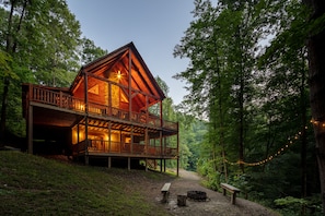 A wooden cabin with a balcony, surrounded by trees and a gravel path.