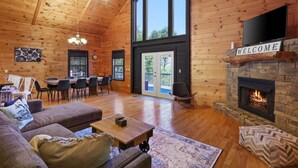 A spacious living room with a fireplace, wooden walls, a dining area, and a large window. - Mountain View Luxury Lodge! Movie Theater, Hot Tub, Fire Pit! (Blue Ridge)