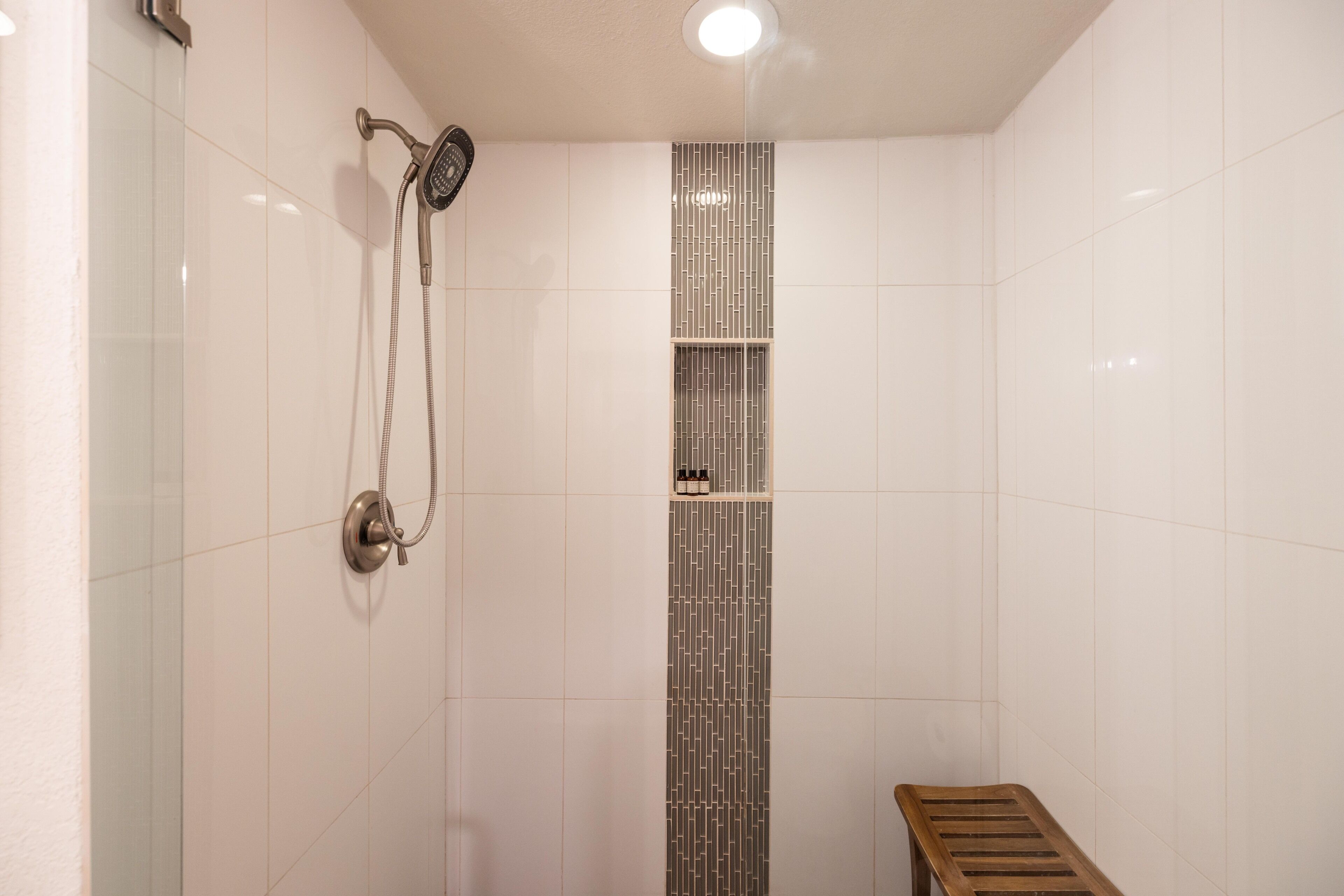 A shower with a handheld showerhead and a wall-mounted faucet, a vertical patterned tile design, and a wooden bench.