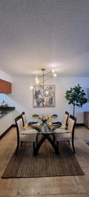 A dining area with a glass table, chairs, a rug, a chandelier, a painting, and a plant.