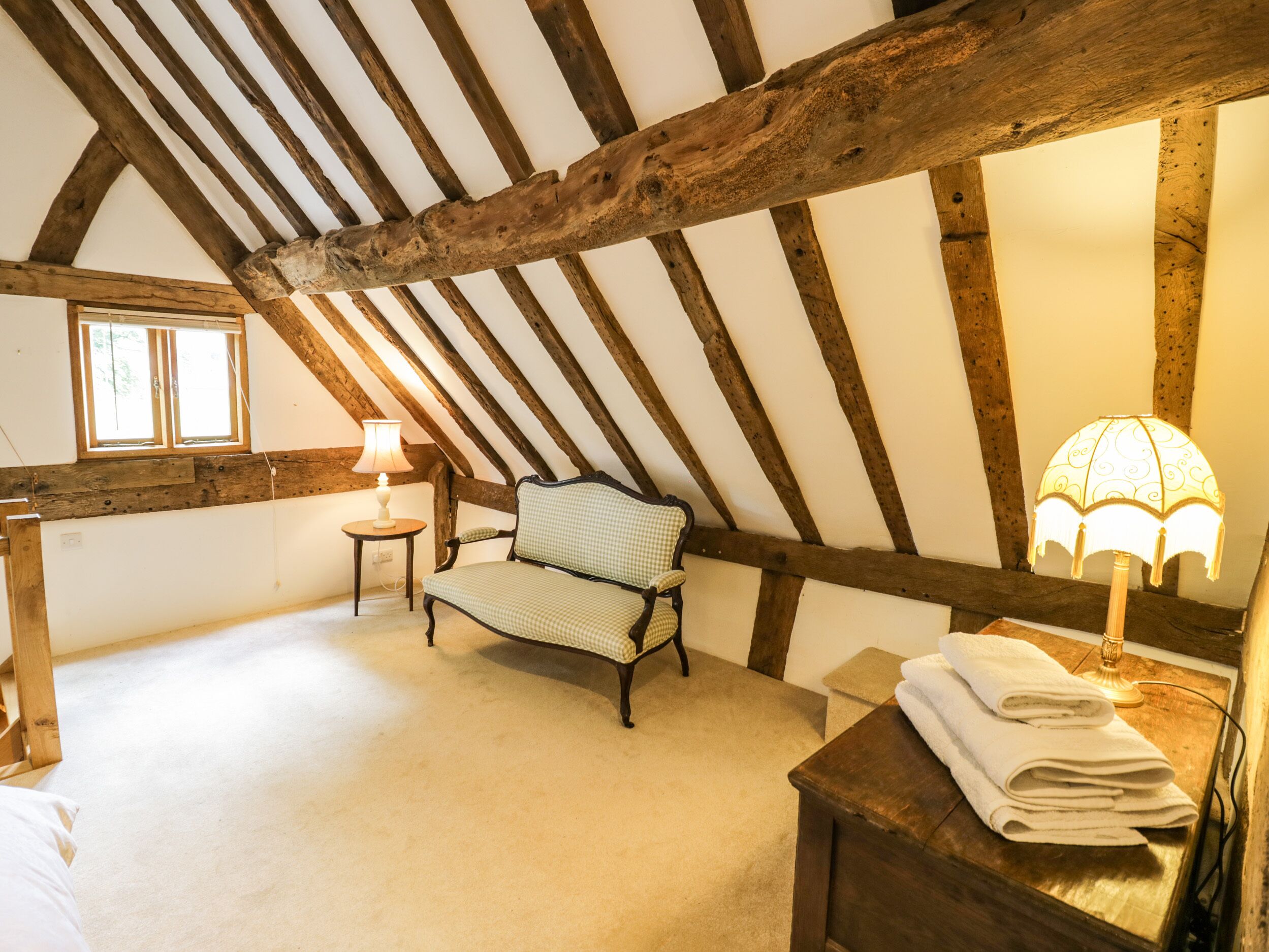 Quarto de sótão acolhedor com vigas de madeira aparentes, uma cadeira xadrez, mesa de cabeceira com abajur e janela.