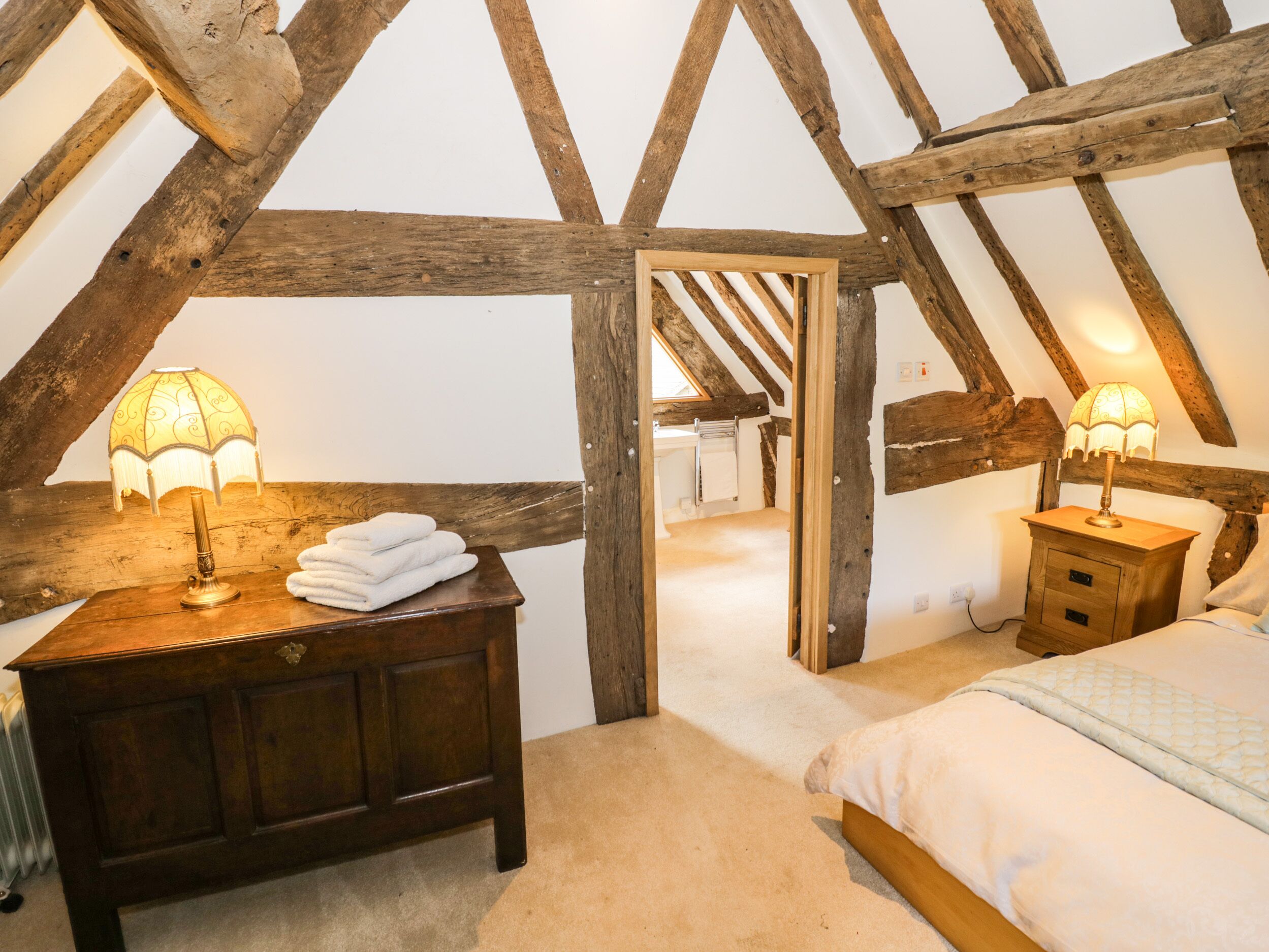 Quarto de sótão aconchegante com vigas de madeira aparentes, um penteou de madeira com uma luminária, uma cama com colcha e vista para um banheiro adjacente.
