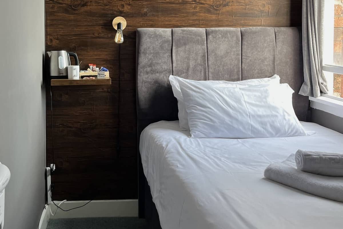 Hotellihuone, jossa on puinen sängynpääty, valkoisilla lakanoilla varustettu sänky, pieni hylly, jossa on hygieniatarvikkeita, ja ikkuna, jossa on verhot.
