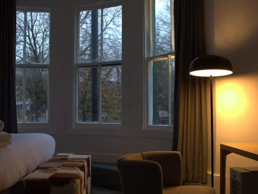 Une chambre d’hôtel avec un lit, une chaise, une table et une lampe. Elle est dotée de trois fenêtres qui laissent entrer la lumière naturelle.