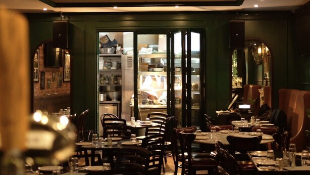 Un ristorante con tavoli apparecchiati per mangiare, una cucina parzialmente visibile tramite vetrate e un'area bar con sgabelli.