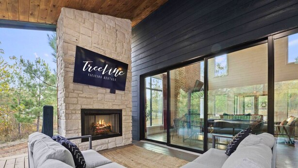 A cozy outdoor seating area with a stone fireplace, a large TV screen, and a glass door leading to a deck.