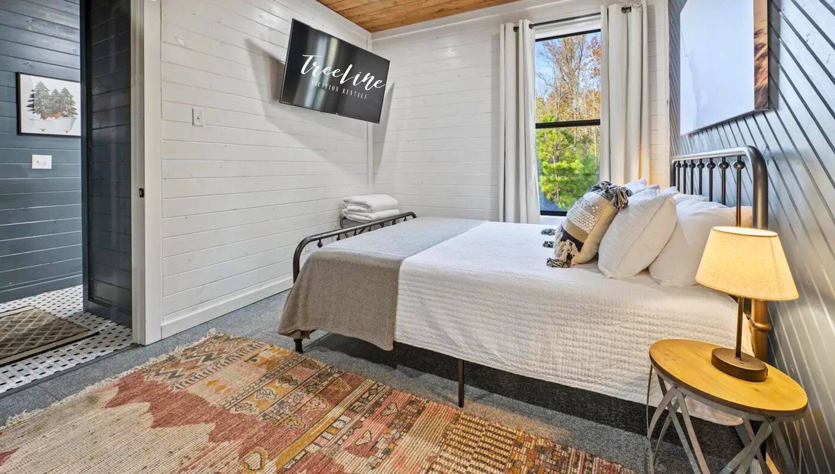 A bedroom with a queen-sized bed, a television mounted on the wall, and a window with a view of trees.