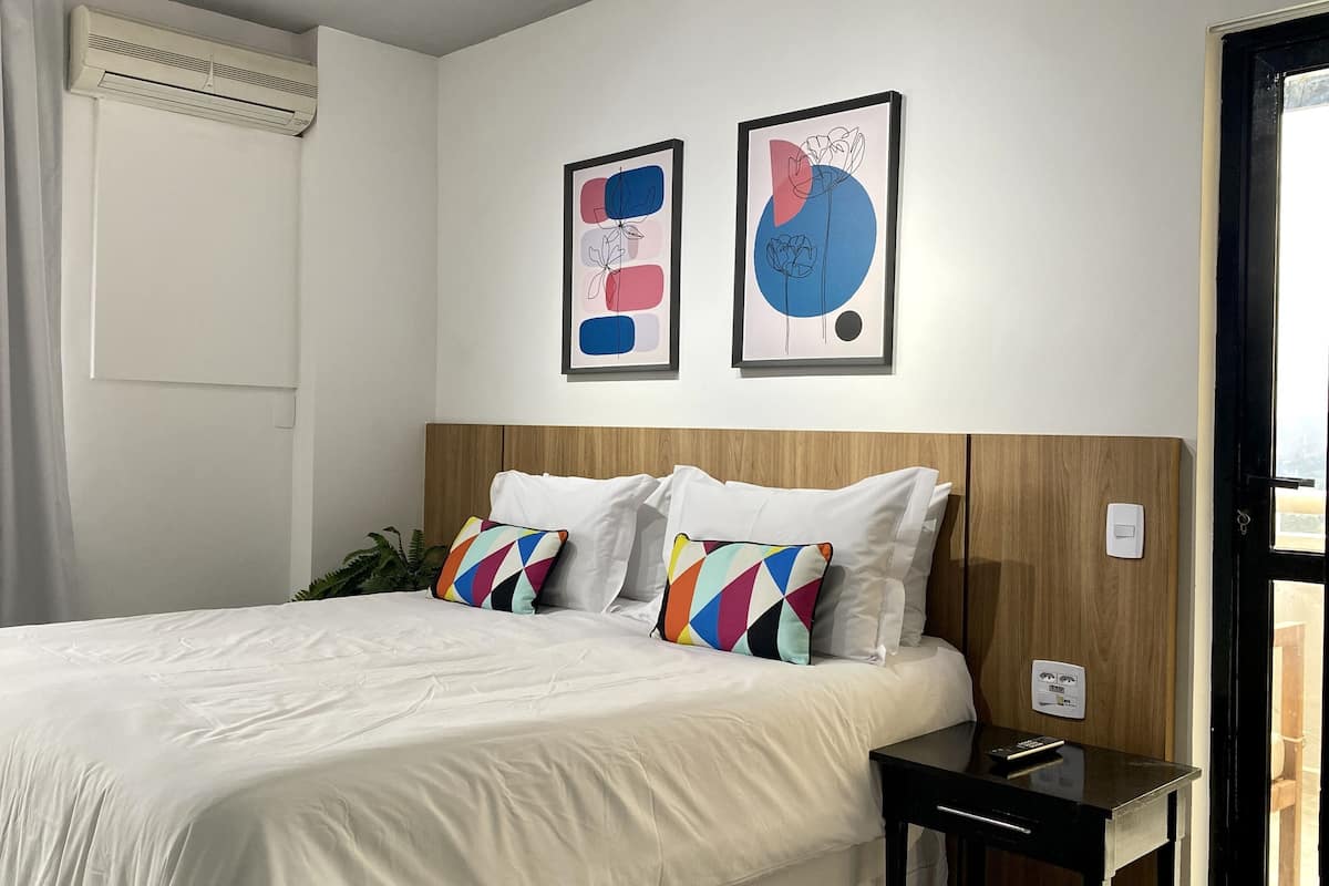 Une chambre d’hôtel comprenant un lit, une table de chevet, un climatiseur fixé au mur et deux œuvres d’art abstraites.