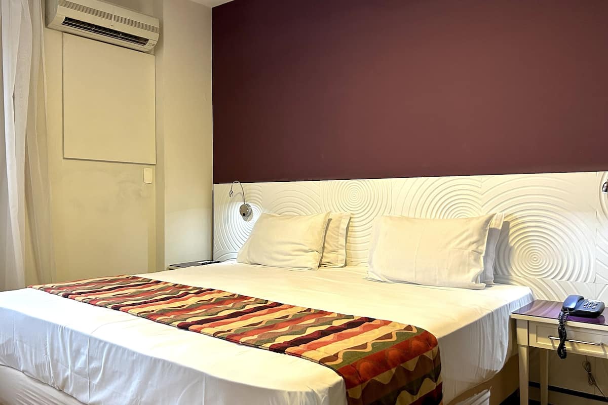 Une chambre d’hôtel comprenant un lit, une table de chevet avec un téléphone, une fenêtre avec des rideaux et un appareil de climatisation.