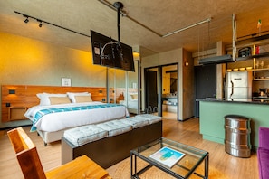 A modern hotel room with a large bed, a sofa, a glass coffee table, and a kitchenette. - QSA - Anora Spaces - Metropolis Alphaville (Barueri)