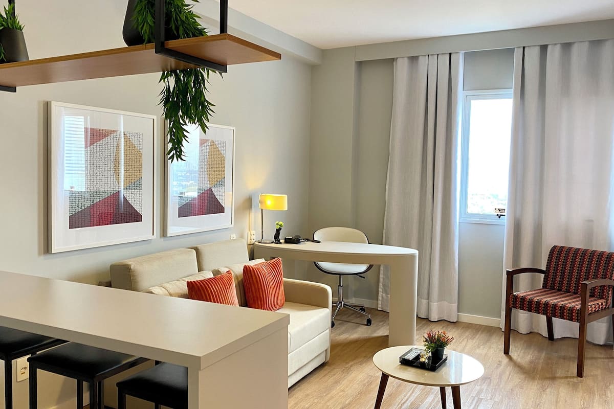 Un salon moderne avec un canapé, une table à manger et une petite table basse. Deux œuvres d’art encadrées ornent le mur, une lampe se trouve sur la table d’appoint et une chaise est agrémentée d’un coussin à motifs.