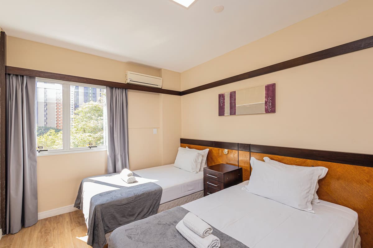 Une chambre d’hôtel avec deux lits, une fenêtre avec des rideaux, un appareil de climatisation, une tête de lit en bois et un miroir fixé au mur.