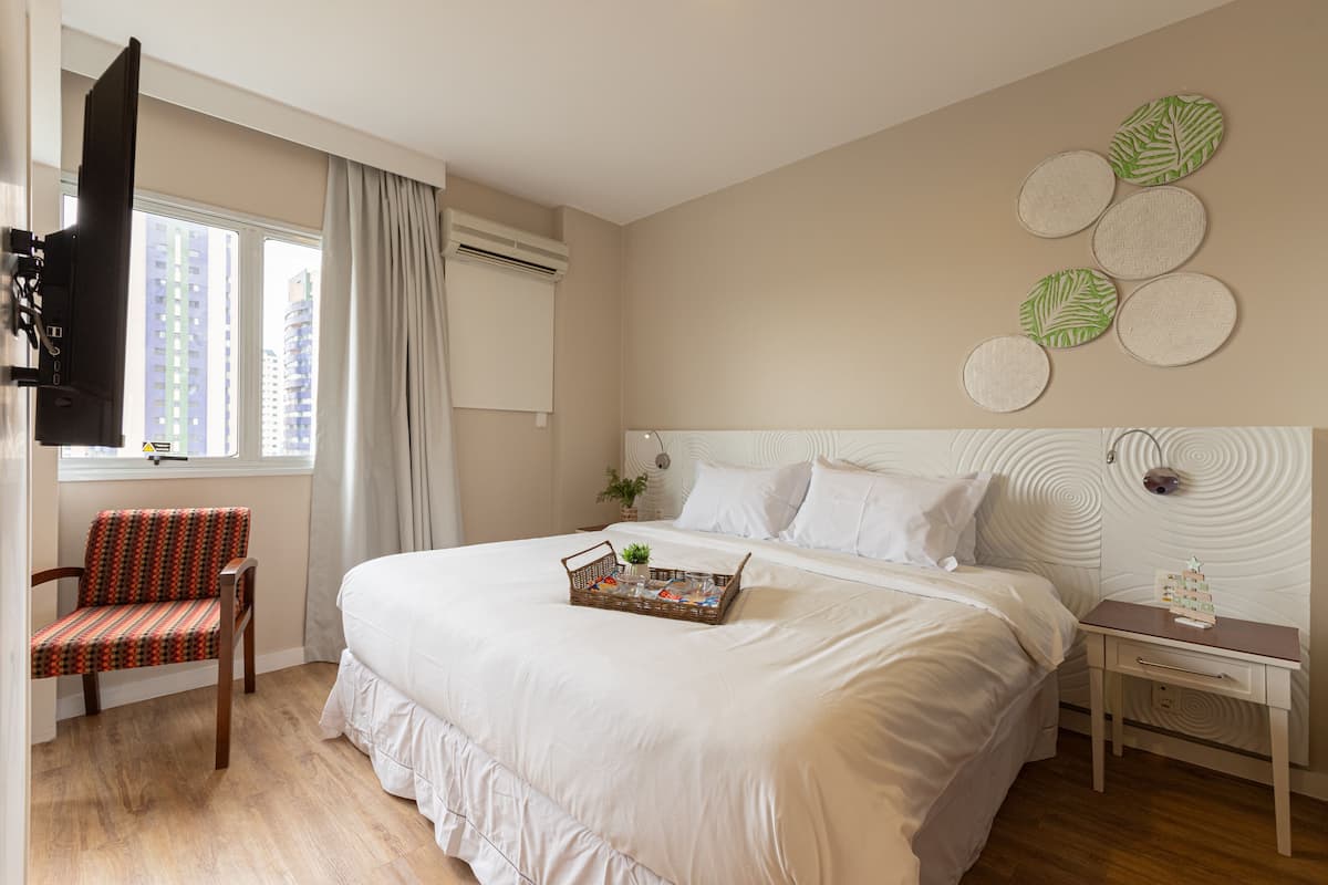 Une chambre à coucher avec un grand lit, une table de chevet avec une horloge, une chaise et une fenêtre avec des rideaux.